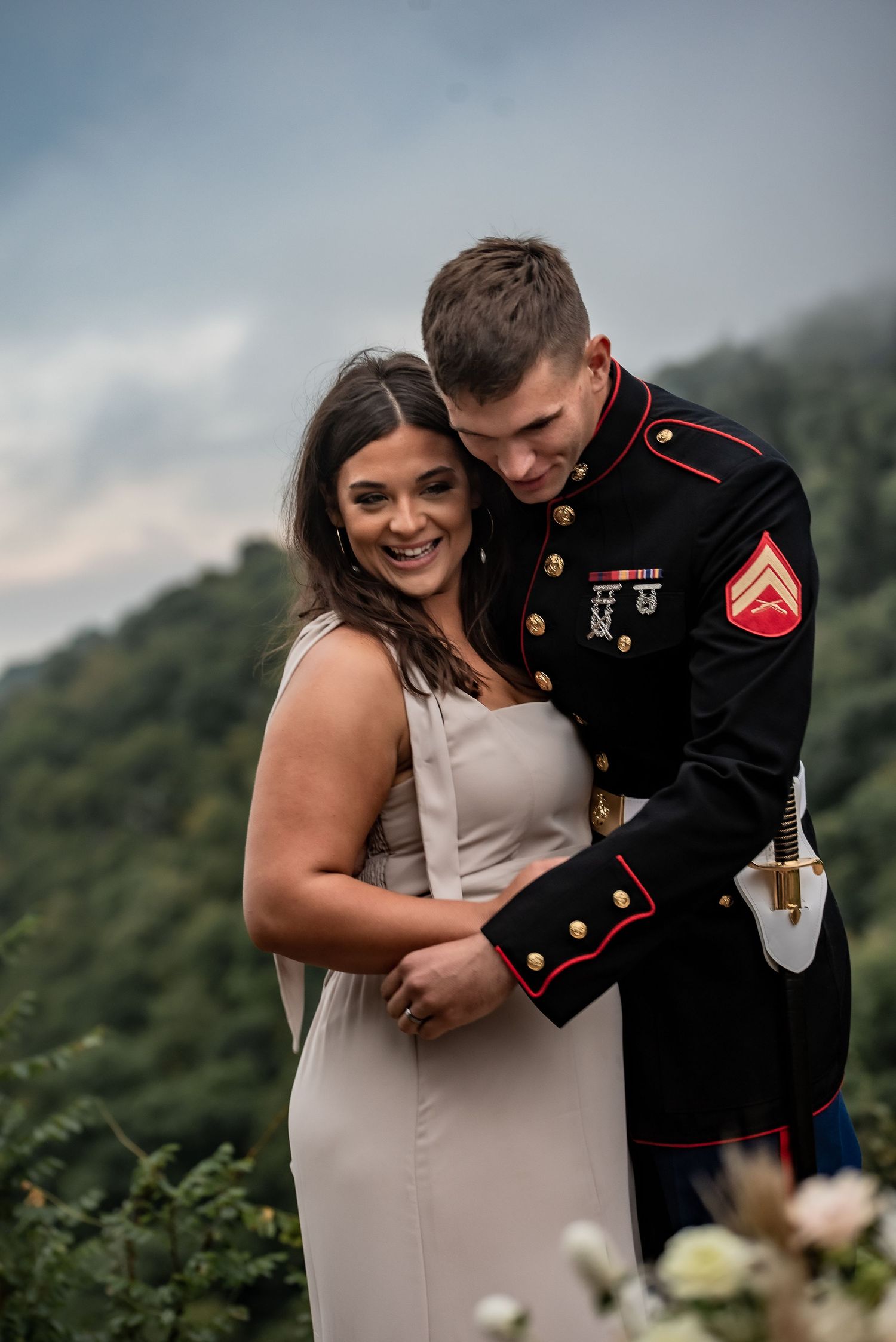 Man in uniform hugs woman as she looks at photographer during mountain elopement session.