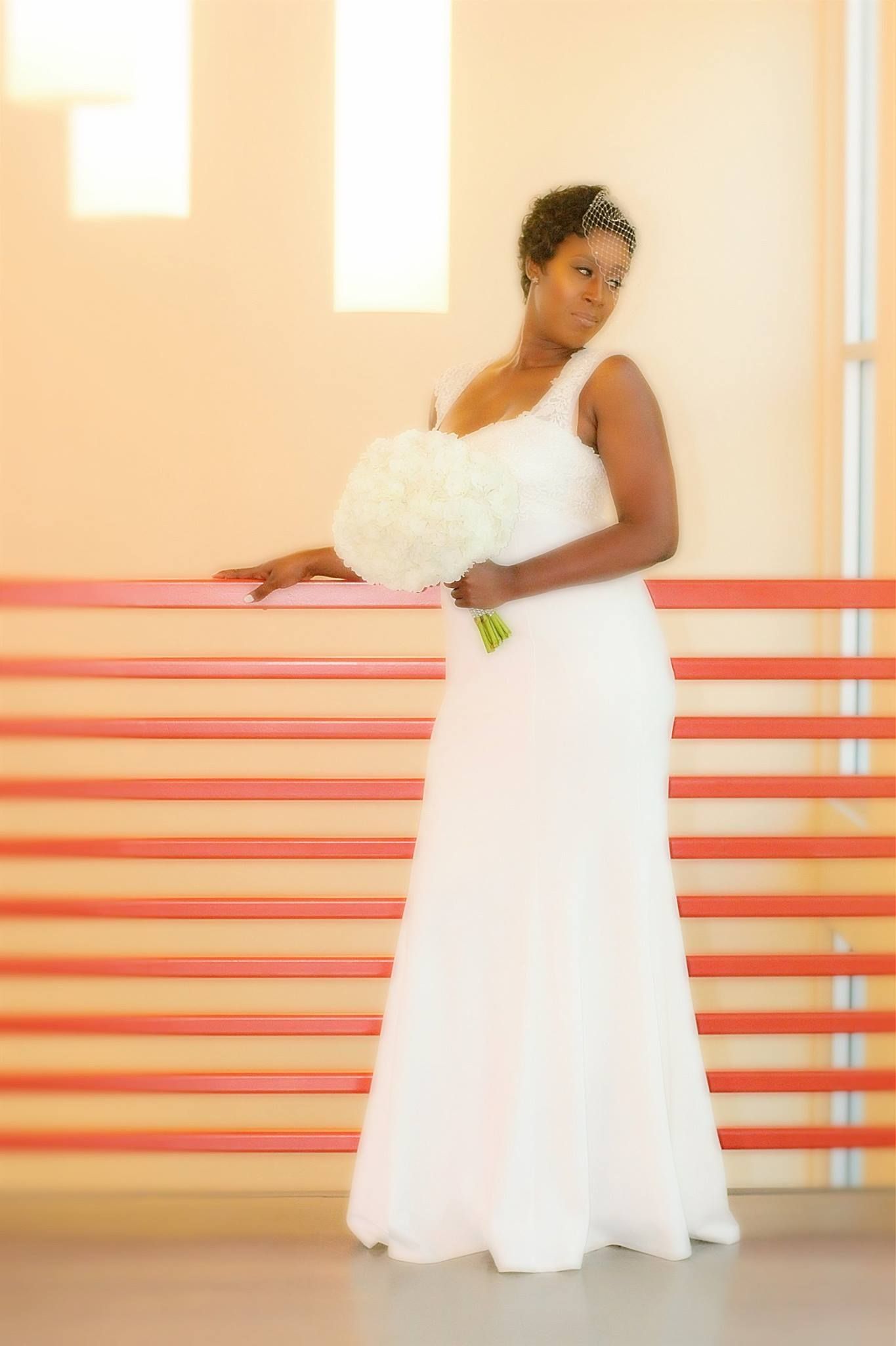 Elegant bride in white flowing gown holding bouquet while posing against orange-striped wall.