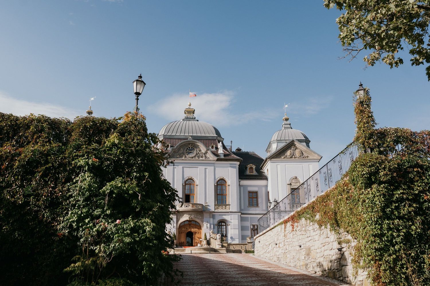 Historic white palace entrance with twin domes and lush garden landscaping.