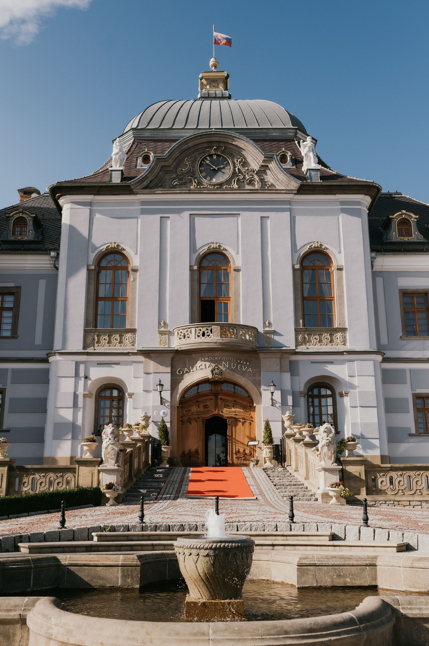 Majestic white palace with domed roof, ornate sculptures and fountain in foreground.