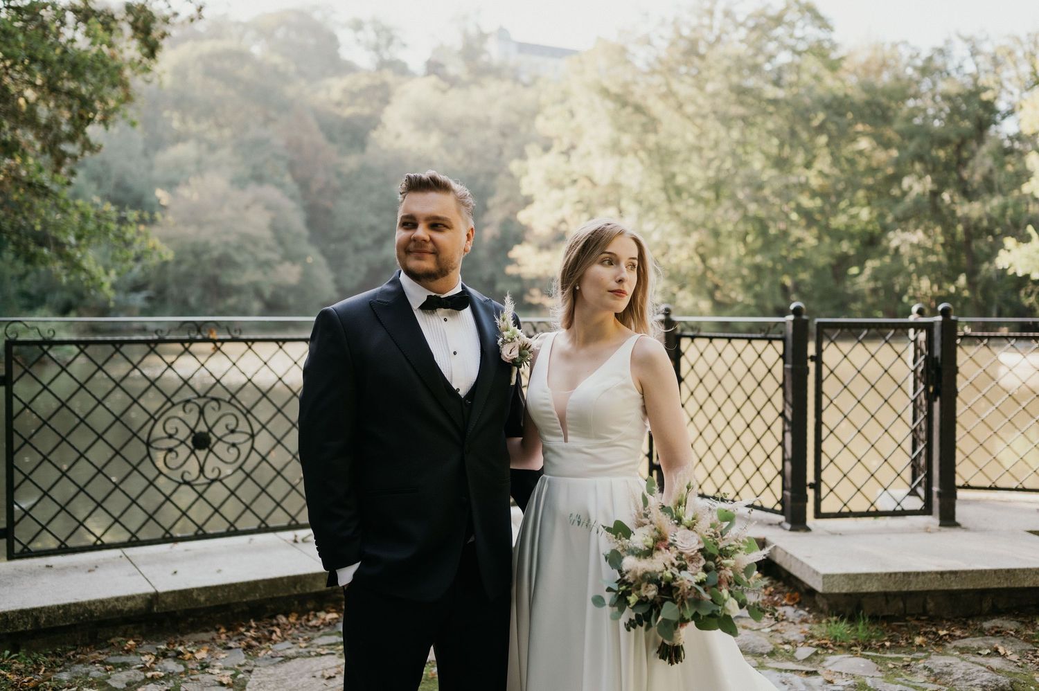 Bride in flowing white dress and groom in black tuxedo pose together on scenic overlook.