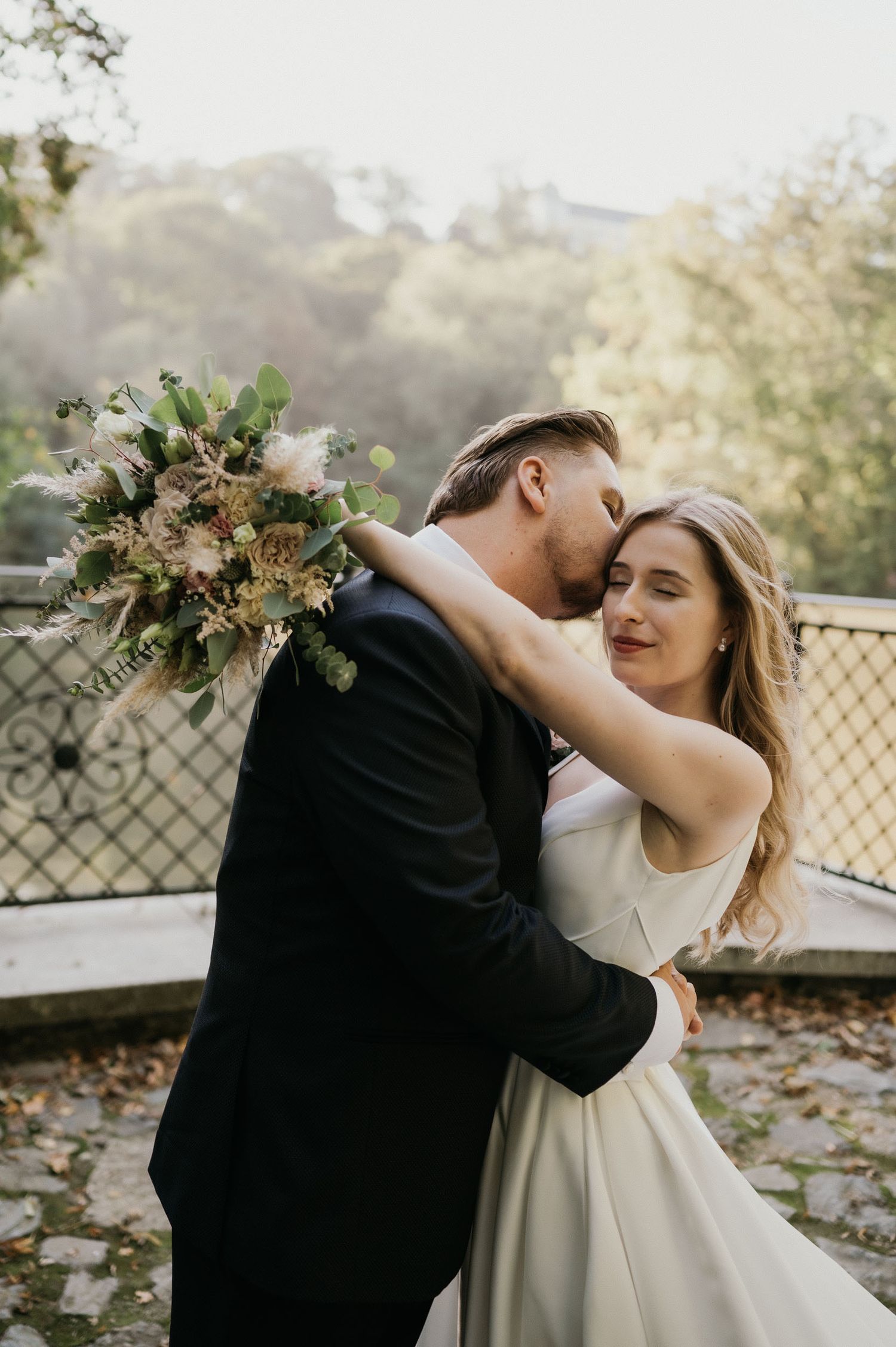 Intimate moment between newlyweds with bride holding bouquet against autumn backdrop.