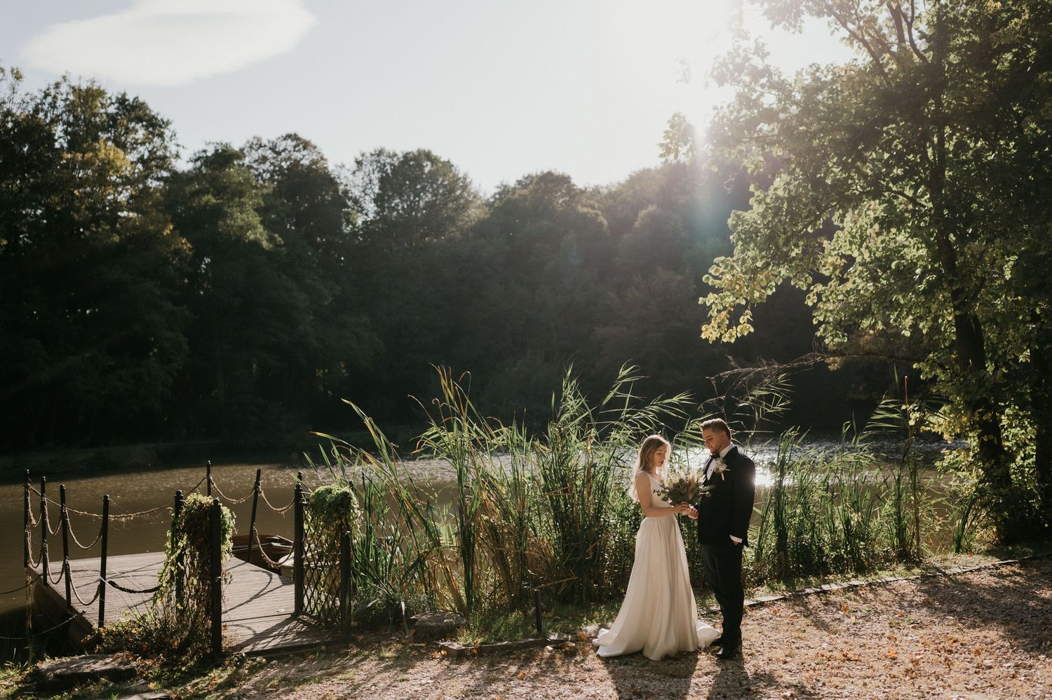 Newlyweds stand together on wooden bridge near tranquil pond with tall grasses.
