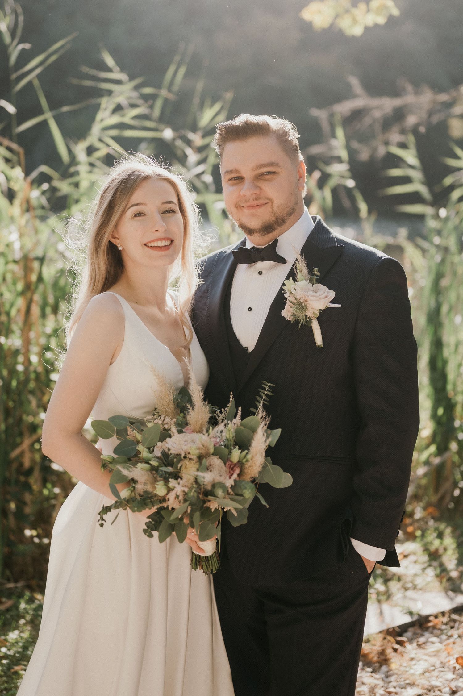 Happy couple in wedding attire pose with bouquet against sun-dappled foliage.