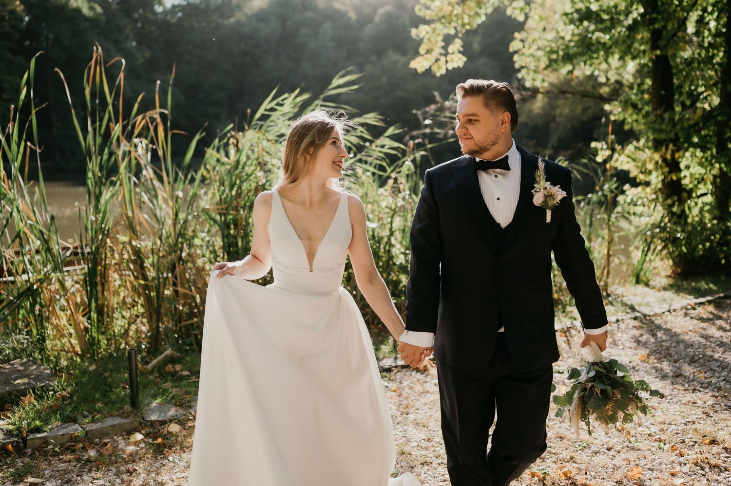 Bride and groom walking hand in hand along sunlit path with natural greenery.