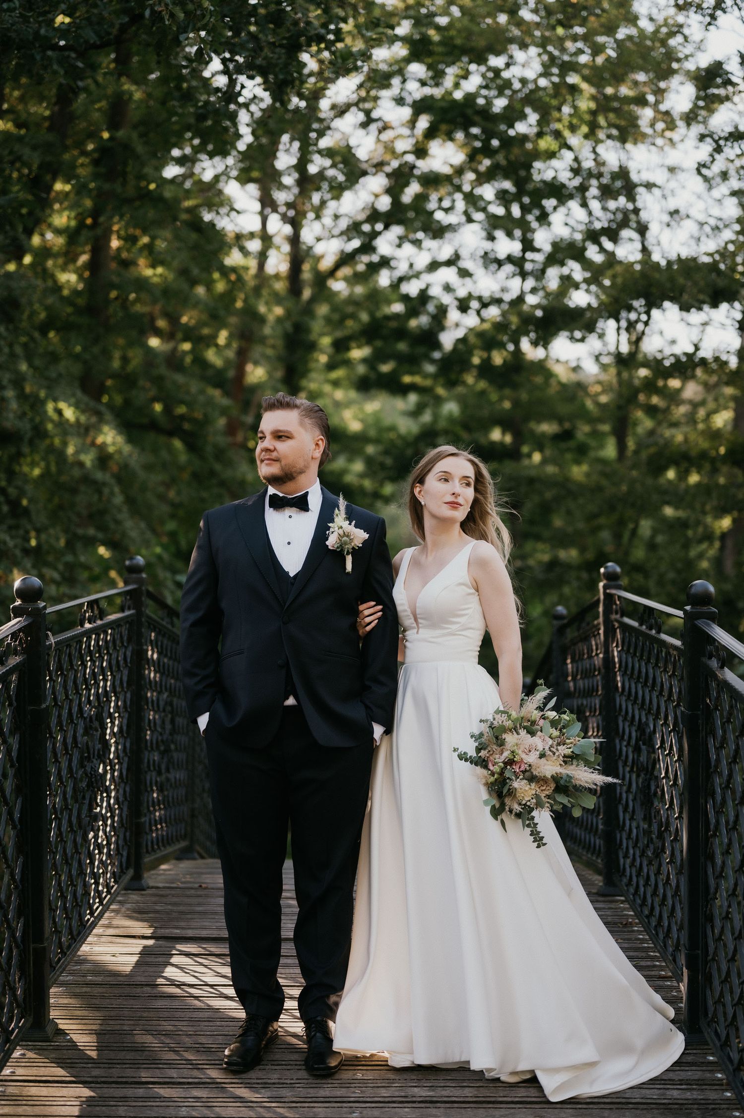 Bride in elegant gown and groom in tuxedo pose on wooden footbridge.