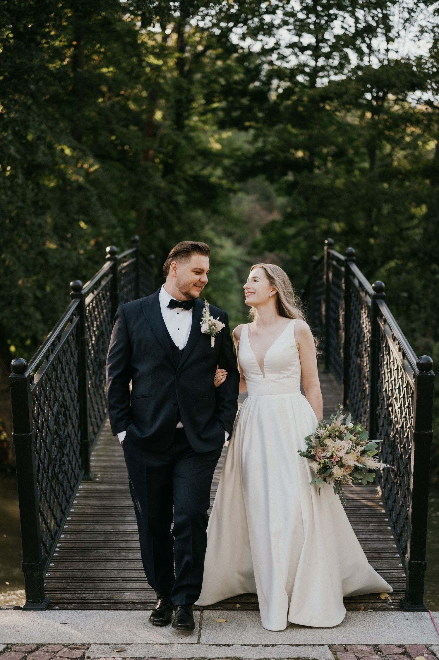 A romantic wedding couple walks arm in arm on an ornate iron bridge surrounded by lush greenery.