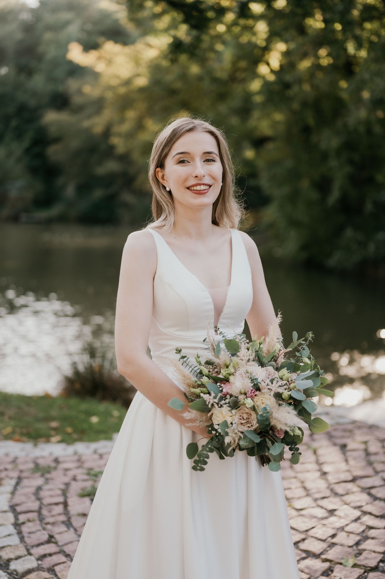 A graceful bride in a plunging v-neck wedding gown holds a soft pink and white bouquet by a river.