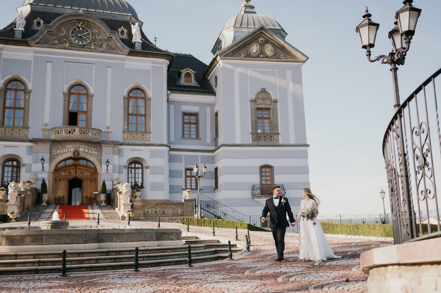 A wedding couple poses in front of a grand white baroque-style palace with ornate architecture.