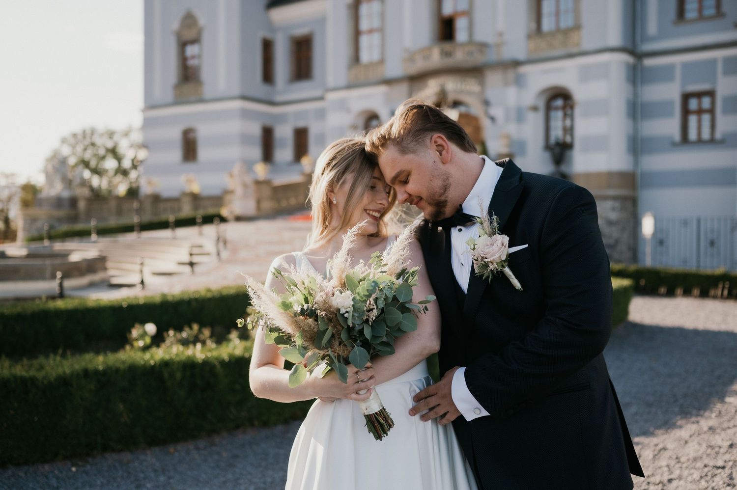 A romantic wedding moment outside a grand palace with lush gardens and ornate architecture.