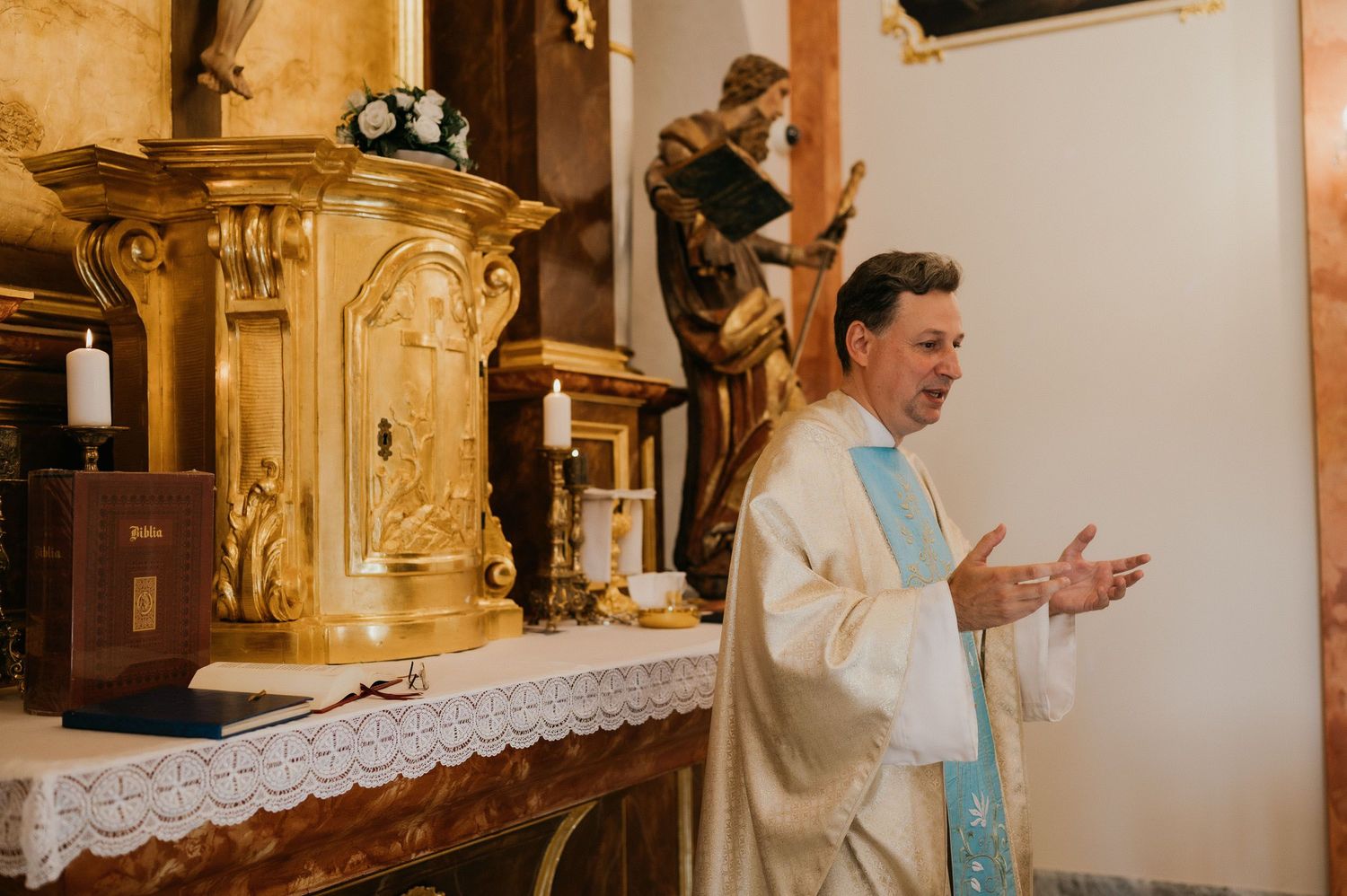 A priest in cream vestments gestures during a Catholic mass service in an ornate church setting.