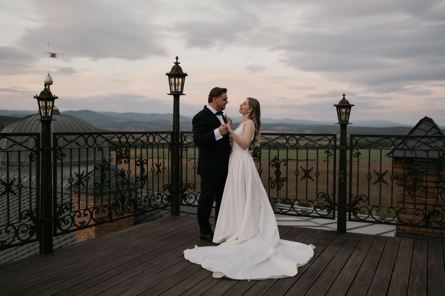 A romantic wedding moment at sunset on an elevated deck with ornate railings and vintage lanterns.
