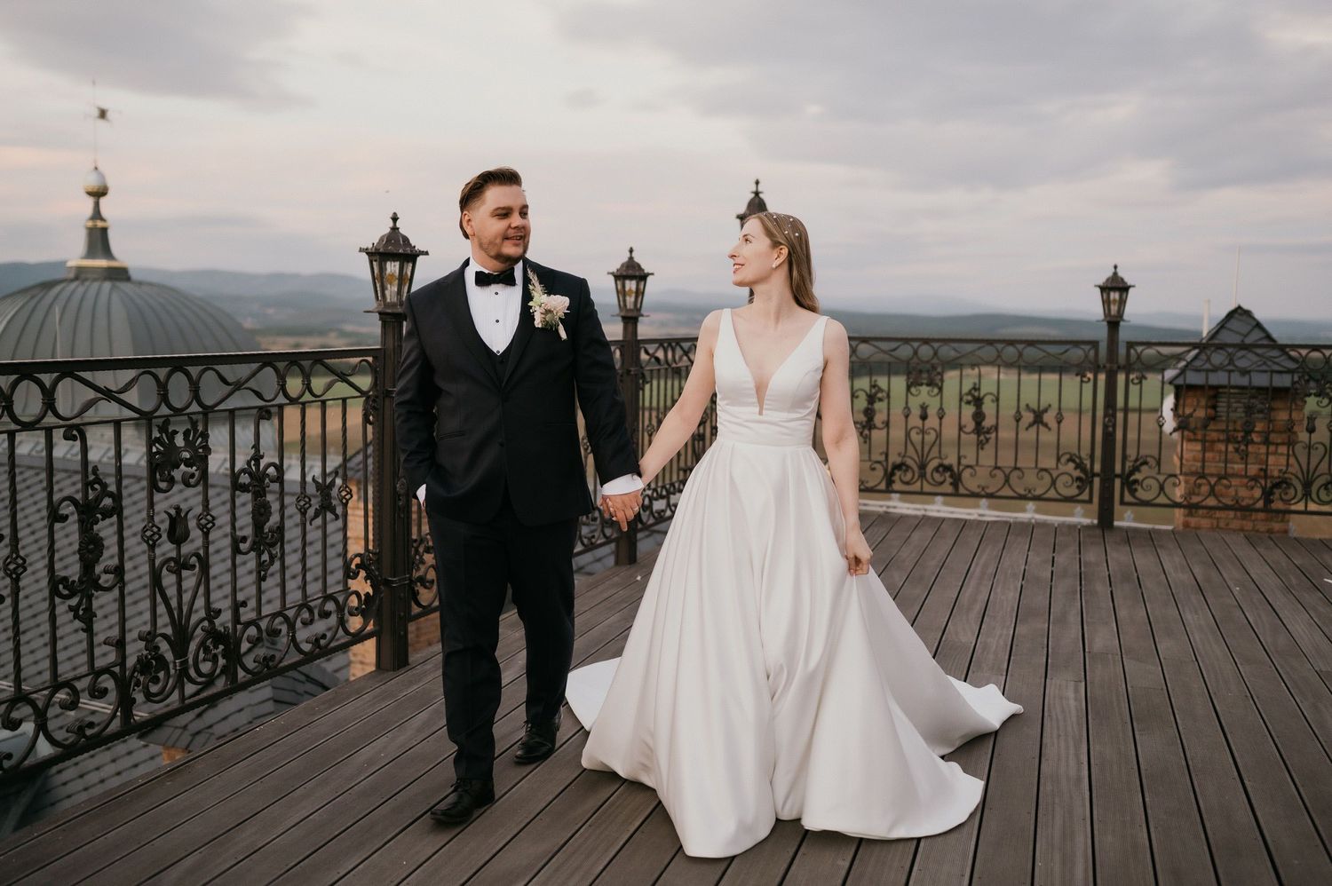 Elegant wedding couple walking on wooden deck with ornate iron railings and mountain views.