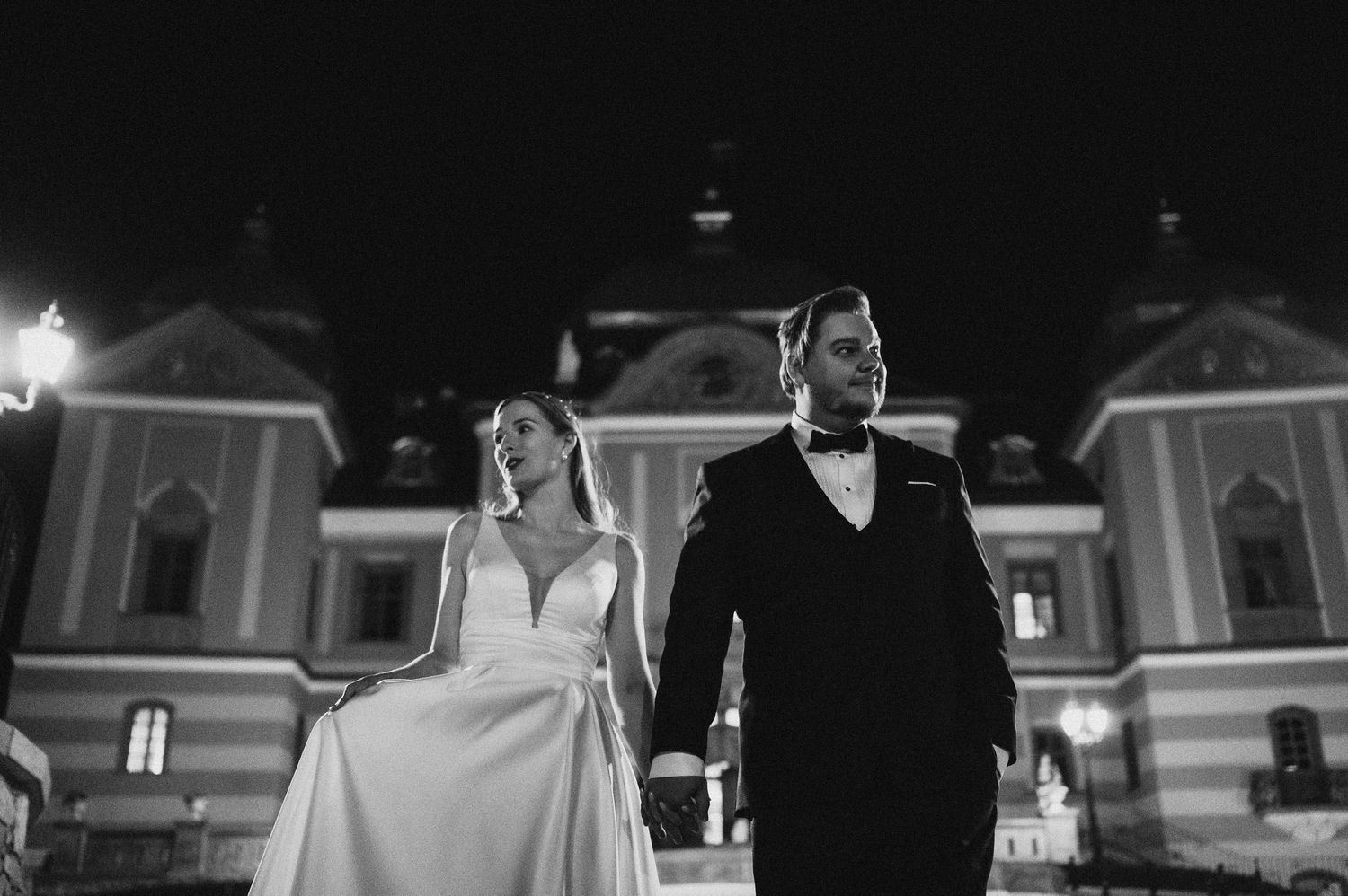 Dramatic low-angle wedding portrait against illuminated historic building at night.