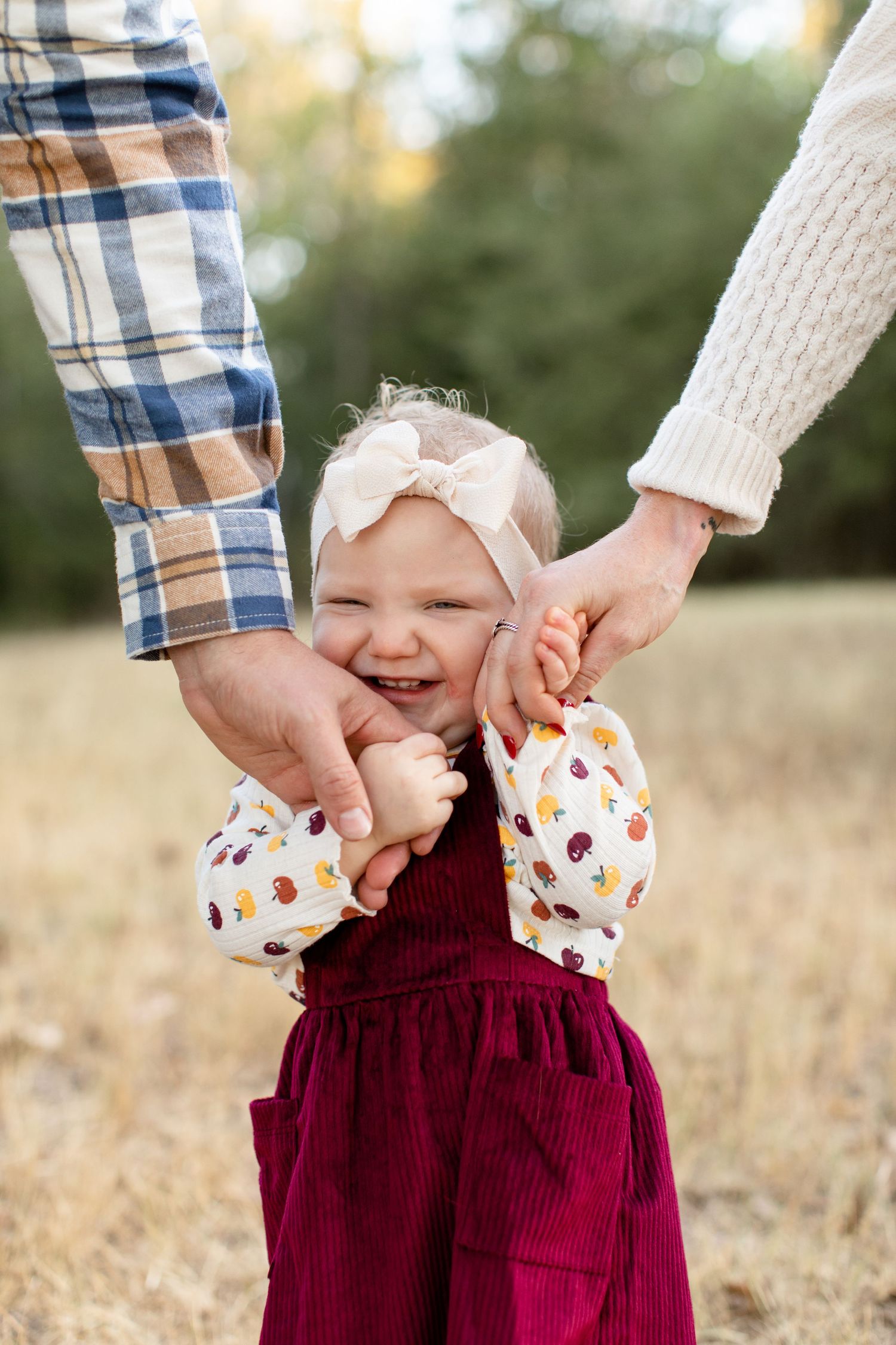 Close-up of laughing toddler in burgundy dress holding parents' hands against natural backdrop.