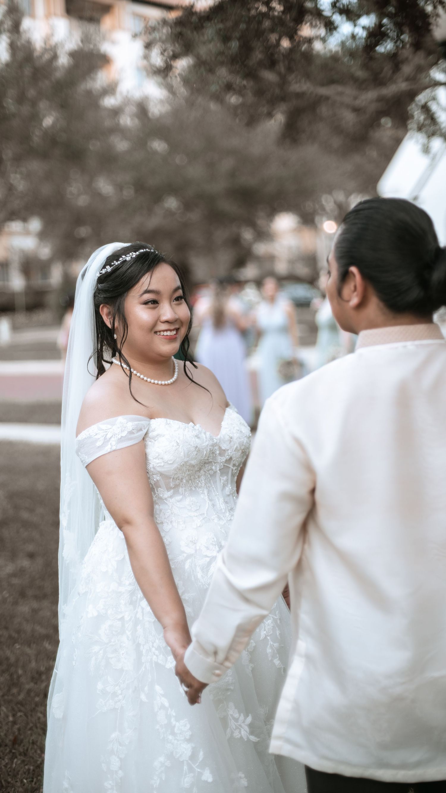 Bride in off-shoulder white lace wedding dress with pearl necklace and tiara holds hands with partner in white barong.