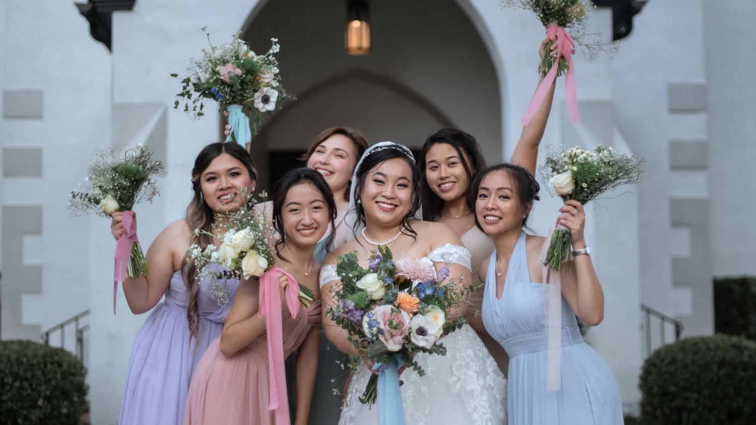 Bride and bridesmaids in pastel dresses pose with bouquets under stone archway.