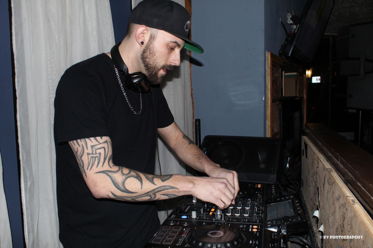 Tattooed DJ in black outfit and cap mixing music on turntables at a nightclub event.
