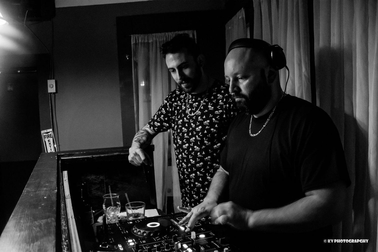 Two male DJs collaborating at a DJ booth in a black and white nightclub photo.