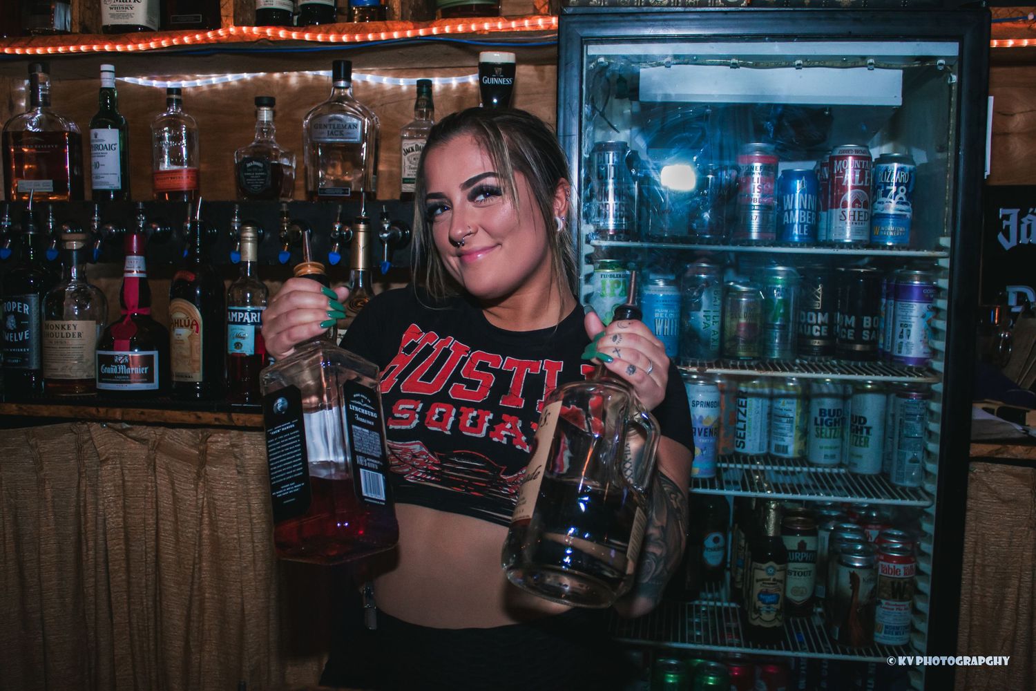 Smiling bartender in Hustle Squad shirt holding liquor bottles in front of a stocked bar.