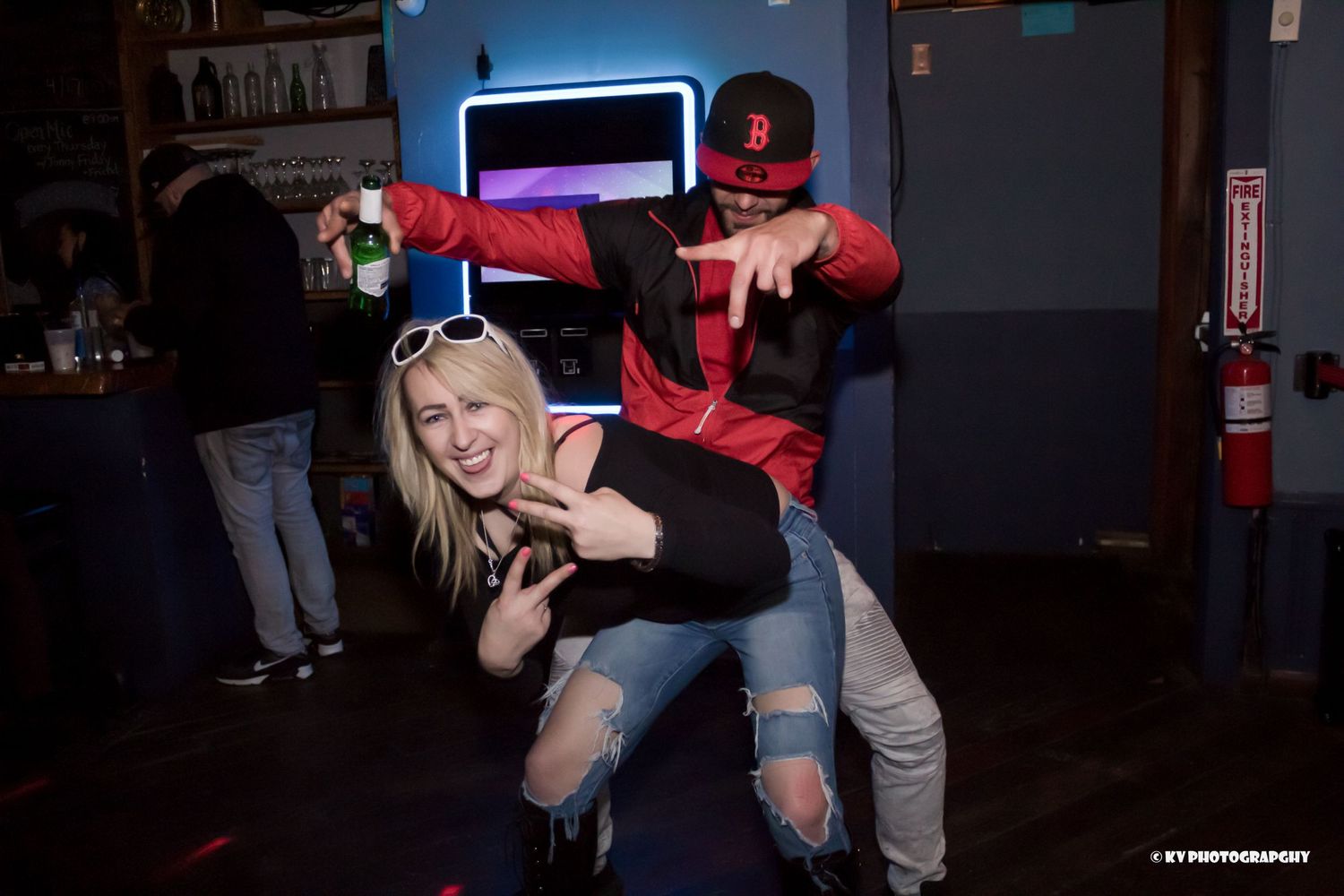 Couple playfully posing at a nightclub, man in red jacket lifting woman in ripped jeans.