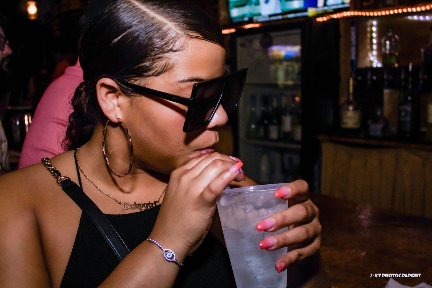 Woman with large sunglasses and pink nails sipping a drink at a bar.