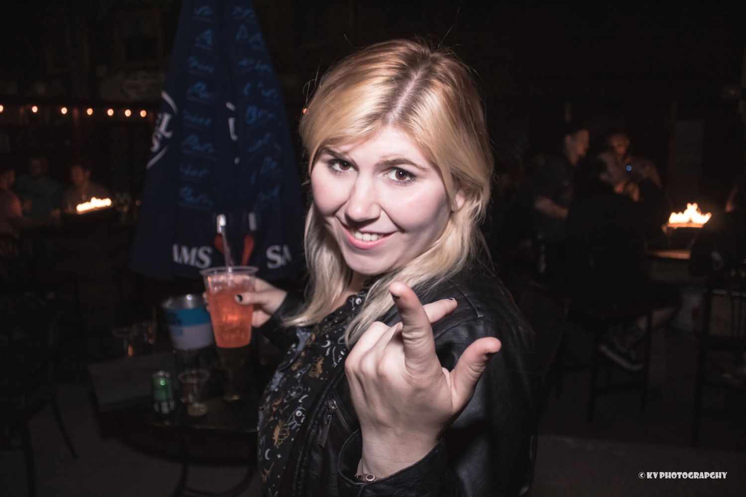 Smiling blonde woman making a rock sign gesture while holding a drink at a bar.