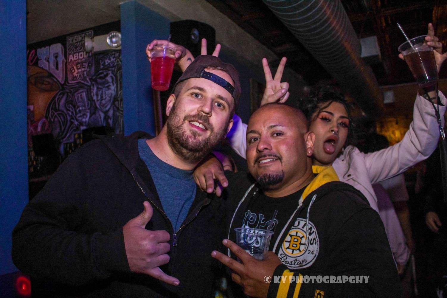 Two smiling men posing together with peace and thumbs-up signs at a lively nightclub.