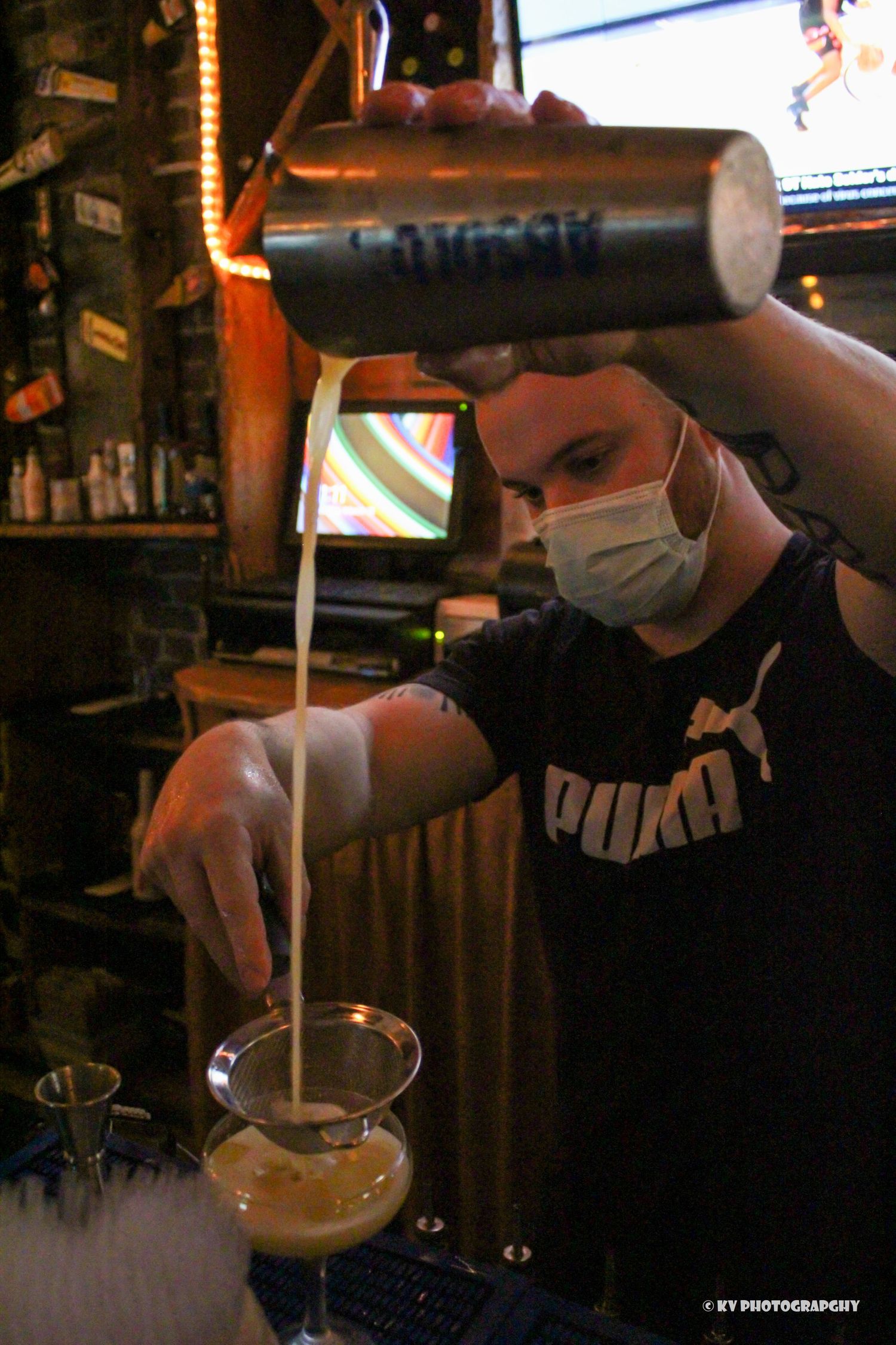Masked bartender pouring cream from a shaker into a cocktail glass at a bar.