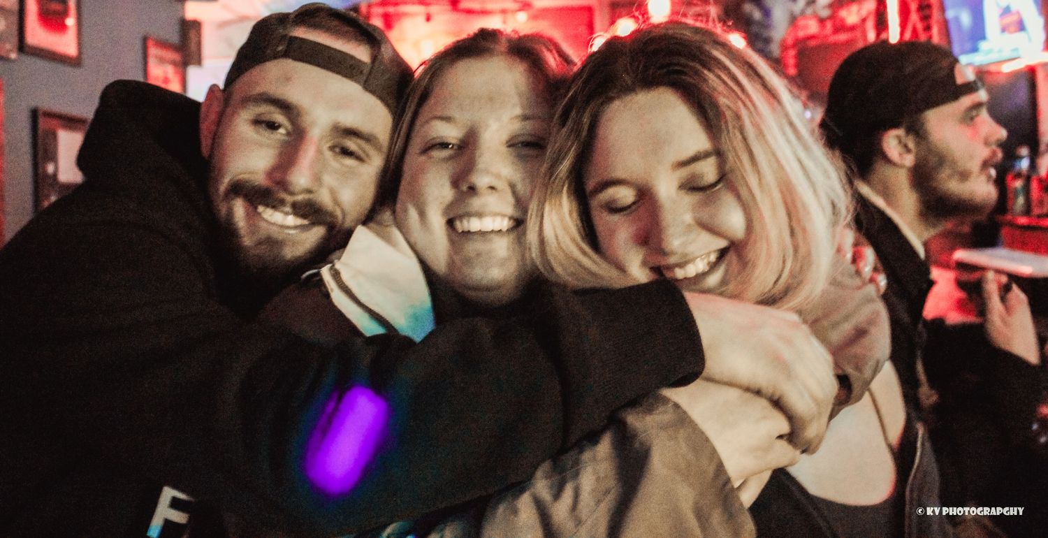 Three friends smiling and hugging together at a vibrant nightclub event.