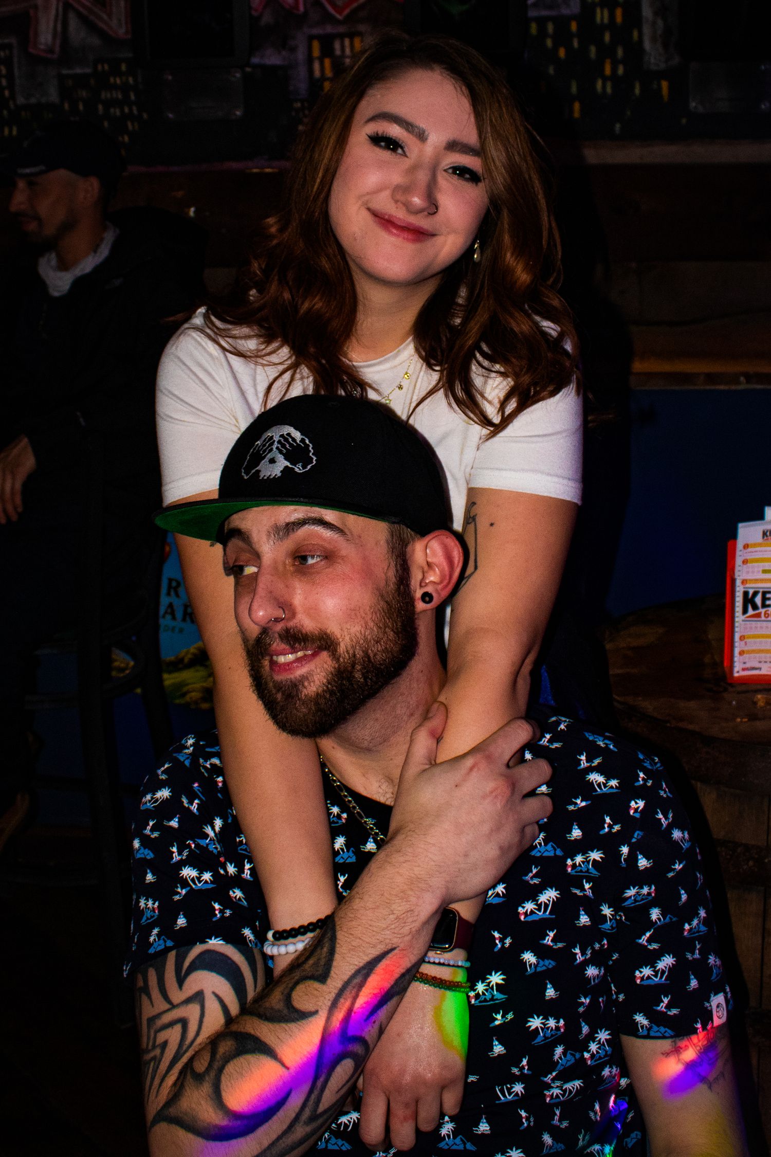 Young woman embracing a seated tattooed man wearing a black cap at a nightclub.