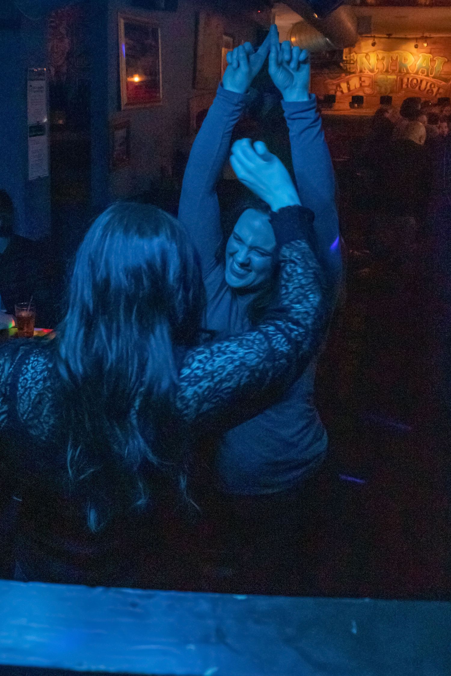 Two women dancing together with raised hands under blue lighting at a bar.