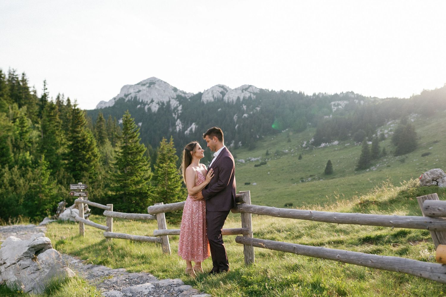 A romantic couple embraces on a wooden fence with majestic mountain peaks and evergreen forest in the background.