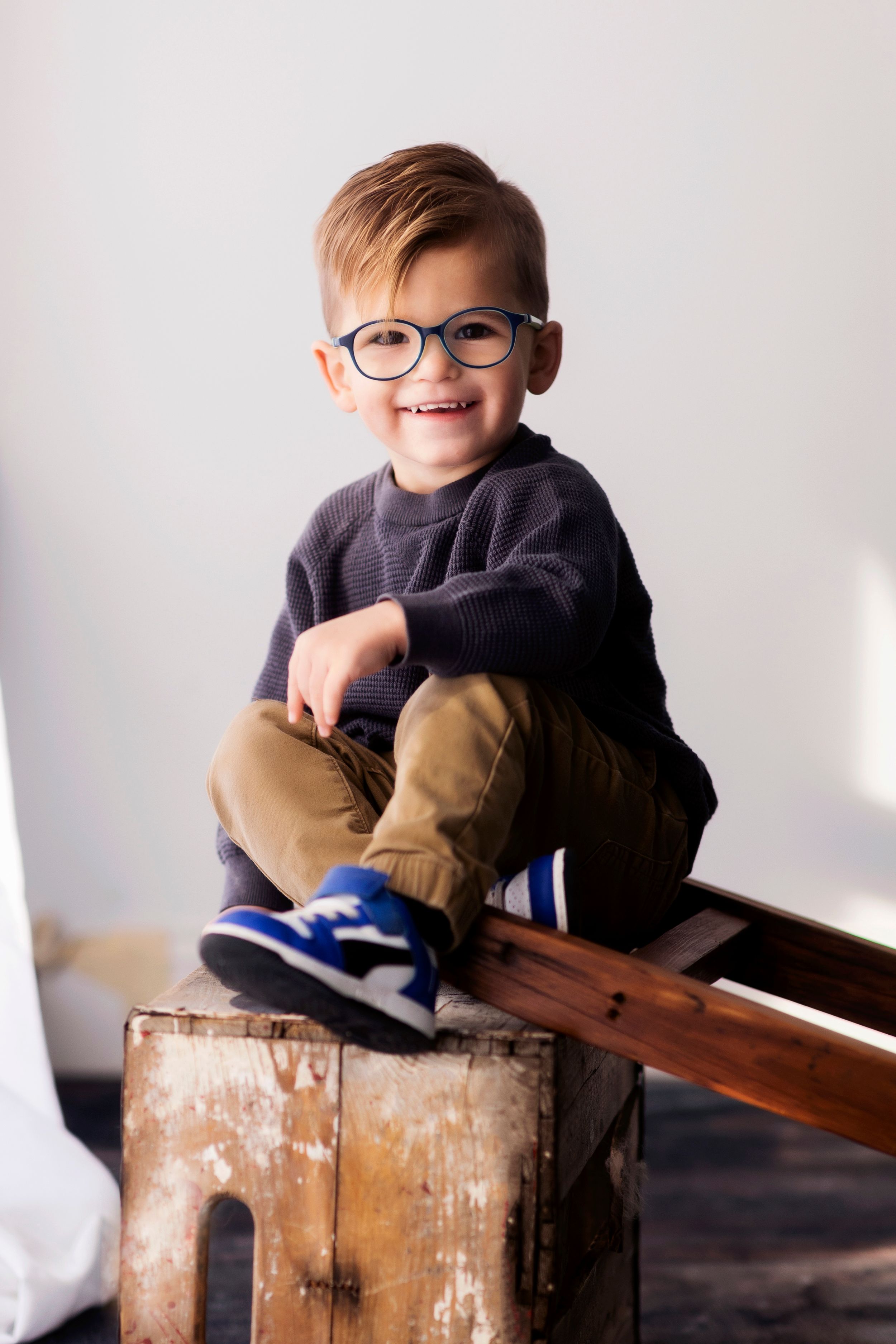 Child wearing glasses and navy sweater posing on rustic wooden crate against white background.