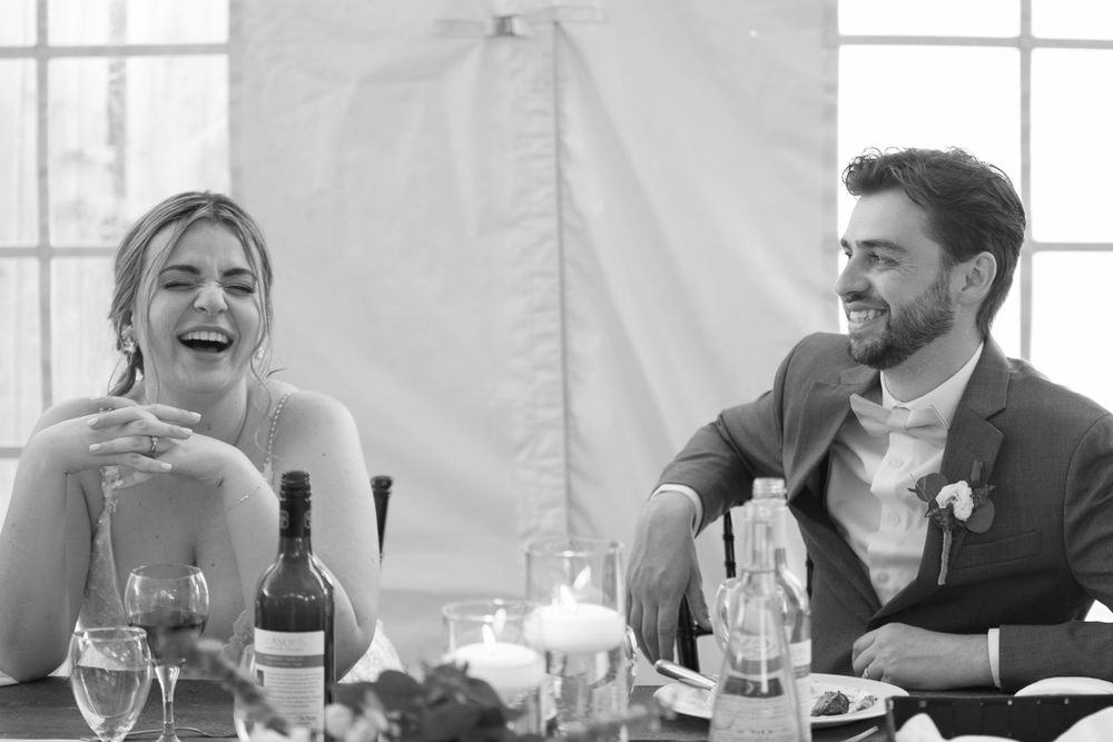 Wedding reception guests laugh and enjoy conversation at a long table indoors.