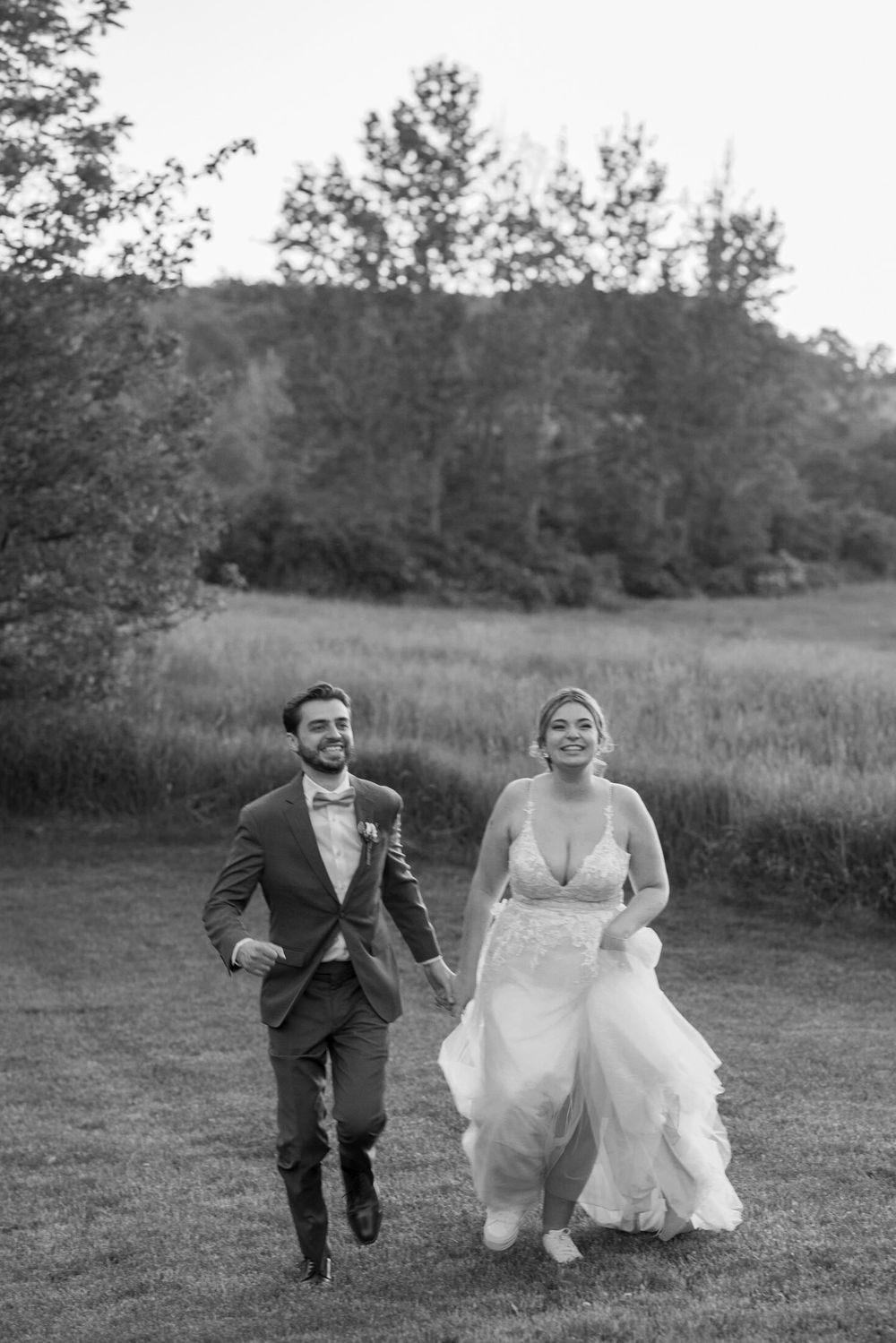 Newlyweds run playfully through a grassy meadow with trees in the background.
