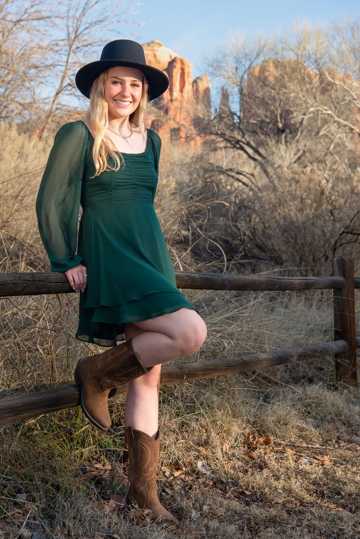 Person in green dress and hat poses at rustic fence with red rock formations in background. Senior Portraits Sedona