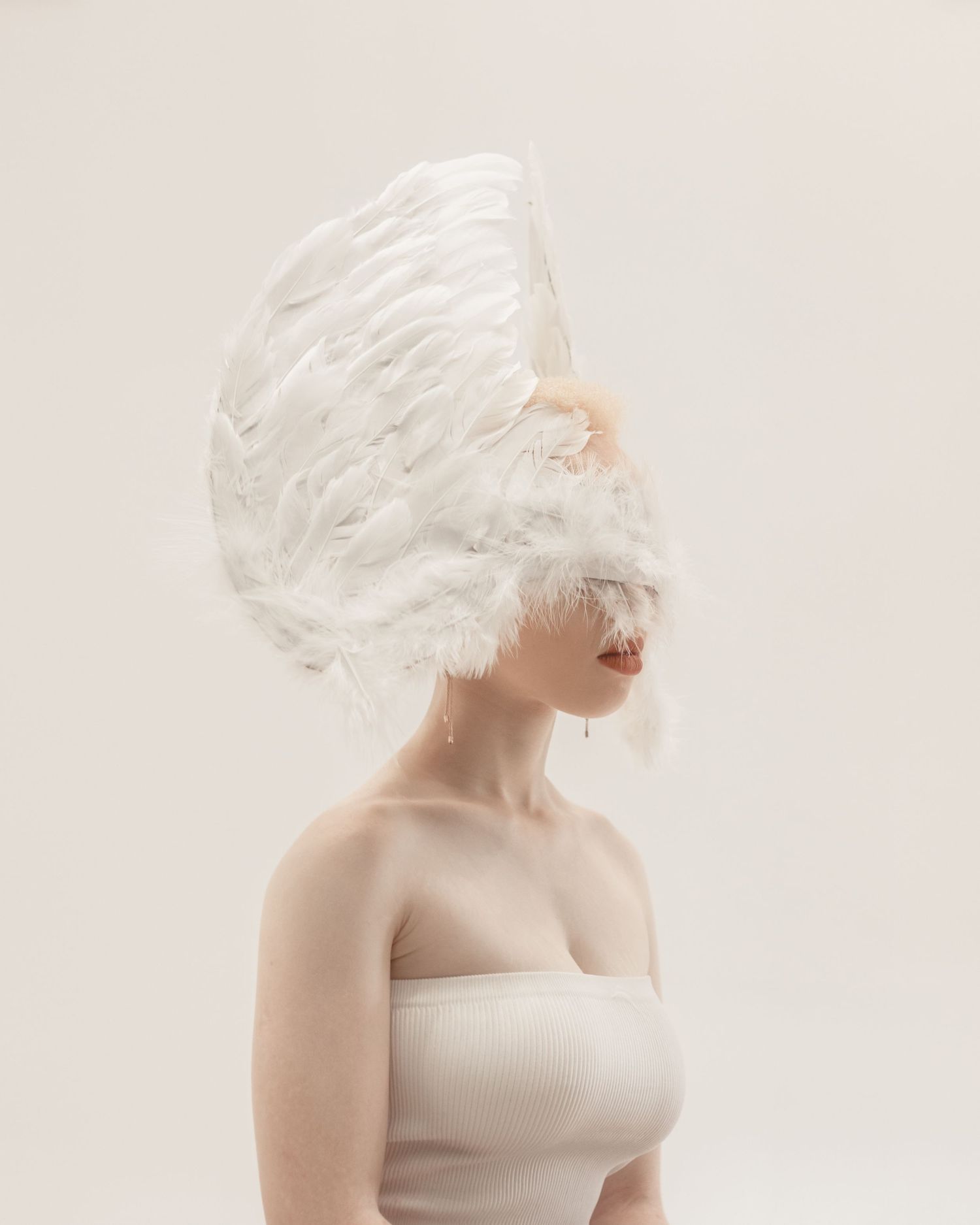 Artistic portrait of someone wearing a white feathered headdress and cream tube top against a light background.