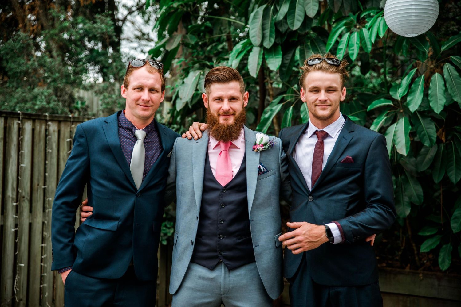 Three stylishly dressed men in suits pose together at a garden wedding.