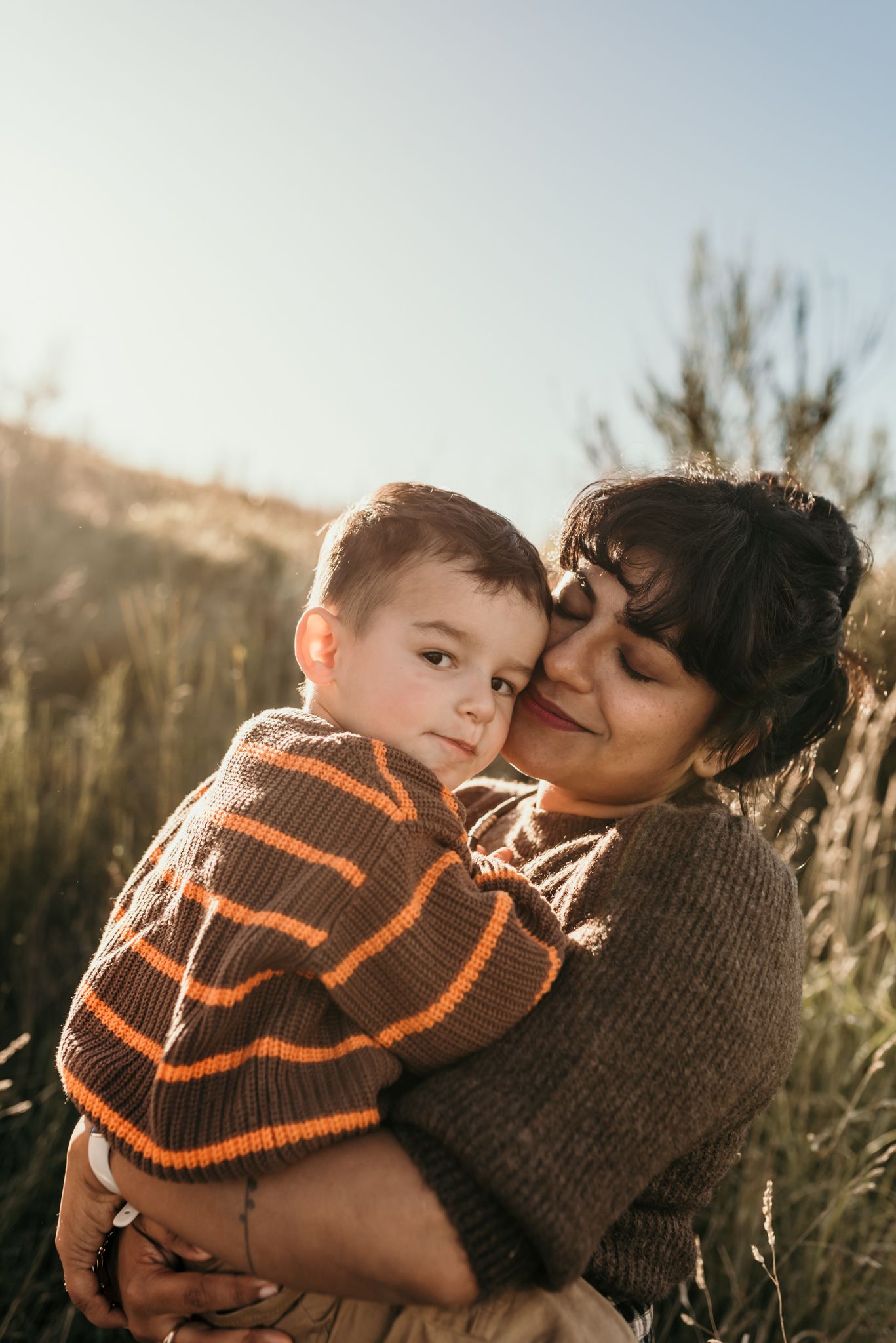 A heartwarming sunset portrait shows a tender embrace between parent and child in striped sweaters among tall grass.