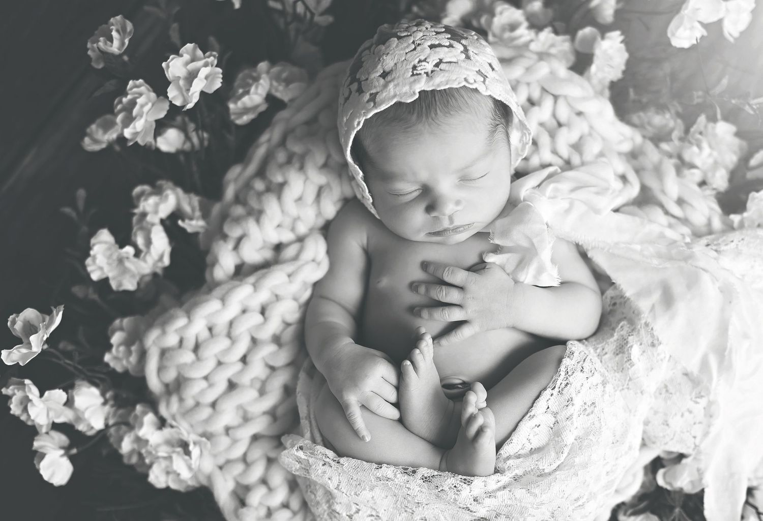 Black and white portrait of a sleeping newborn on textured knit blanket surrounded by flowers.