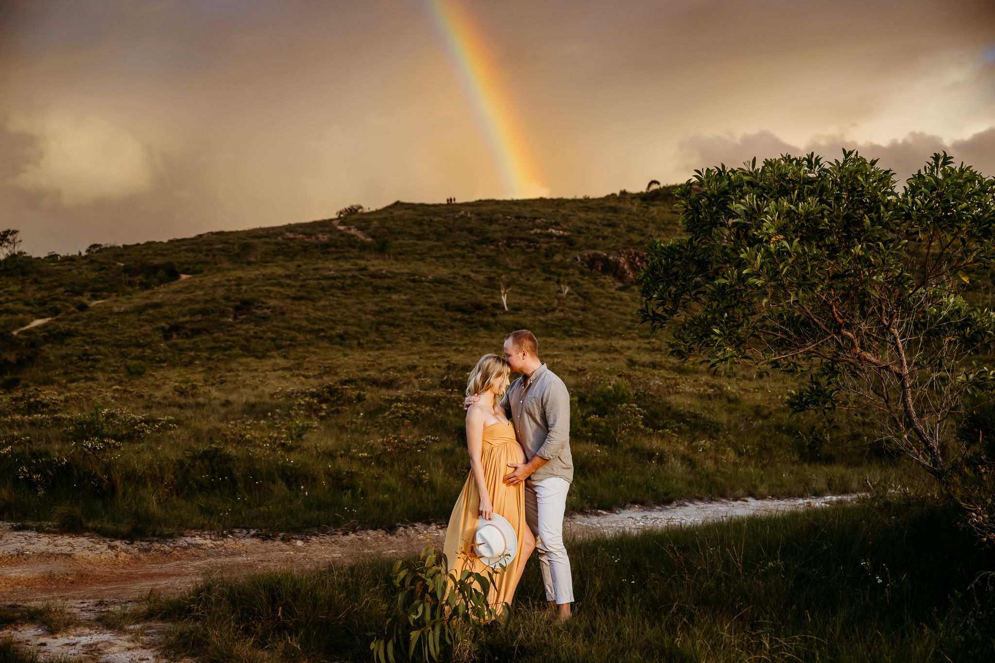 Couple embracing under dramatic rainbow sky on hilltop path.