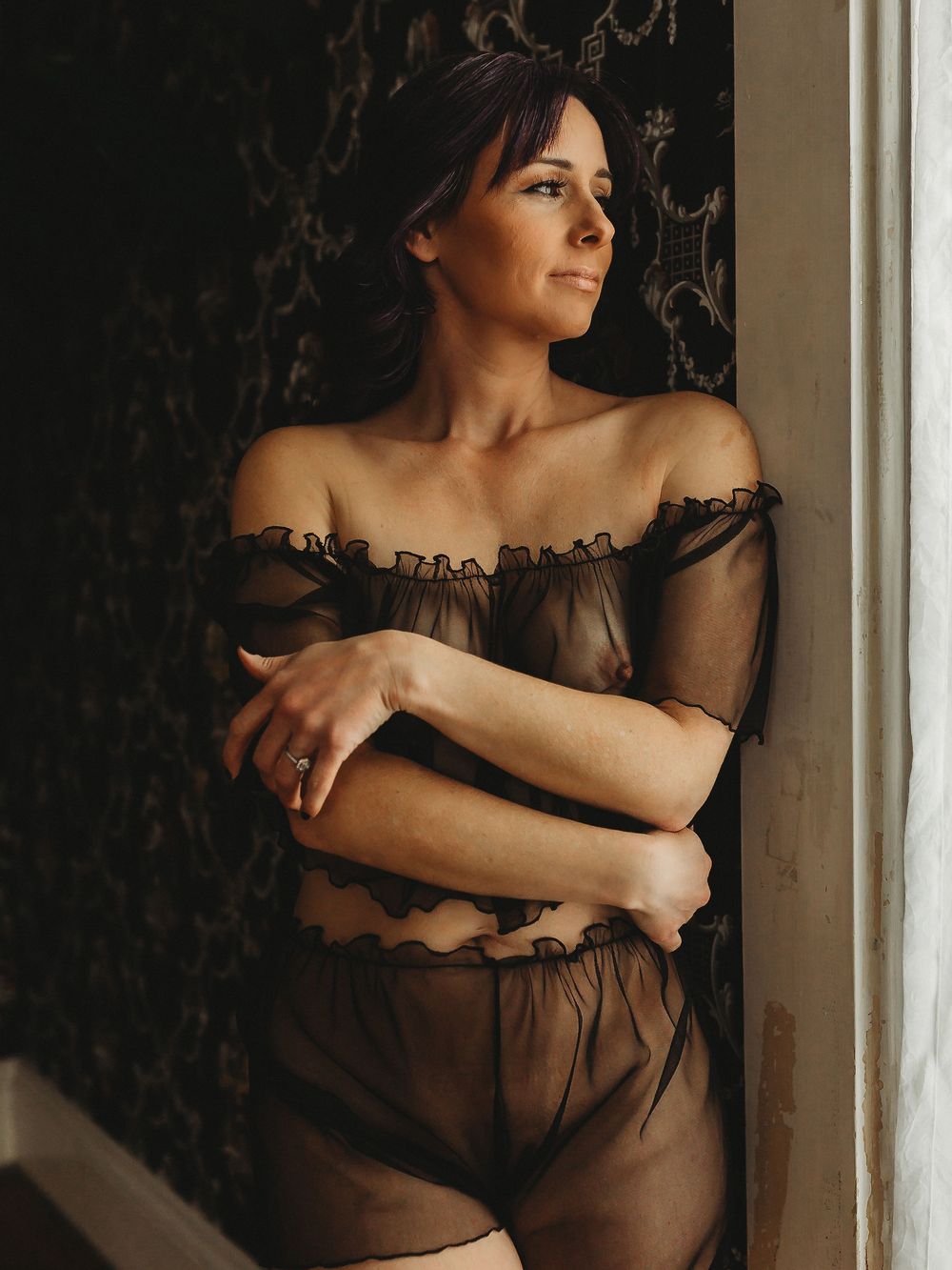 Romantic boudoir photo with off-shoulder sheer dress against vintage wallpaper.