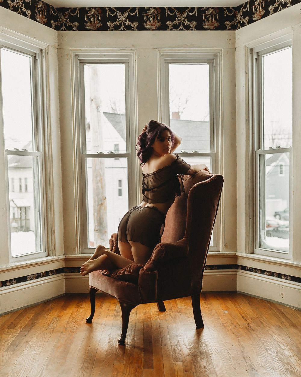 Vintage-style boudoir photo in bay window with velvet chair and natural lighting.