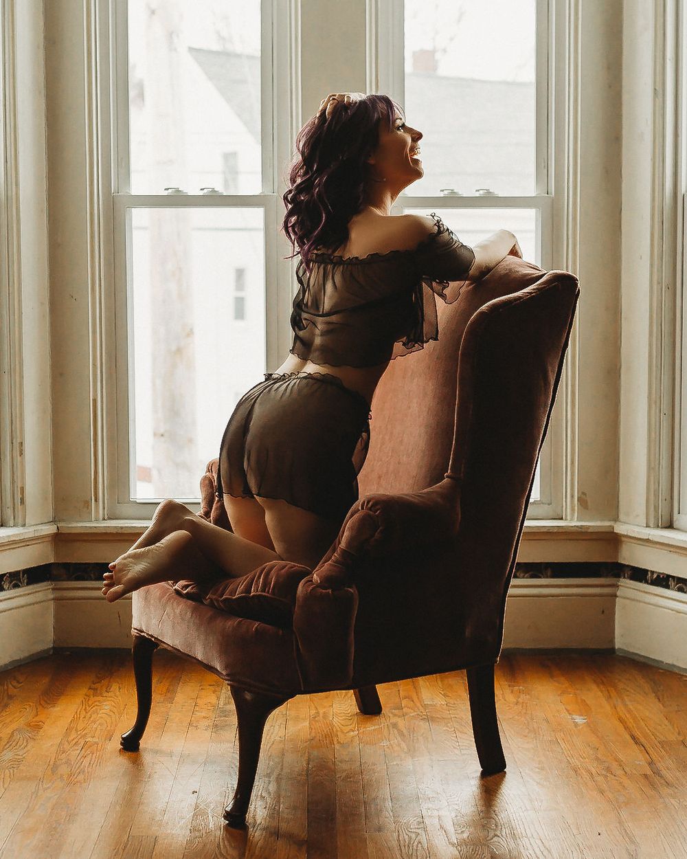 Elegant portrait in brown dress seated in antique chair by bright window.