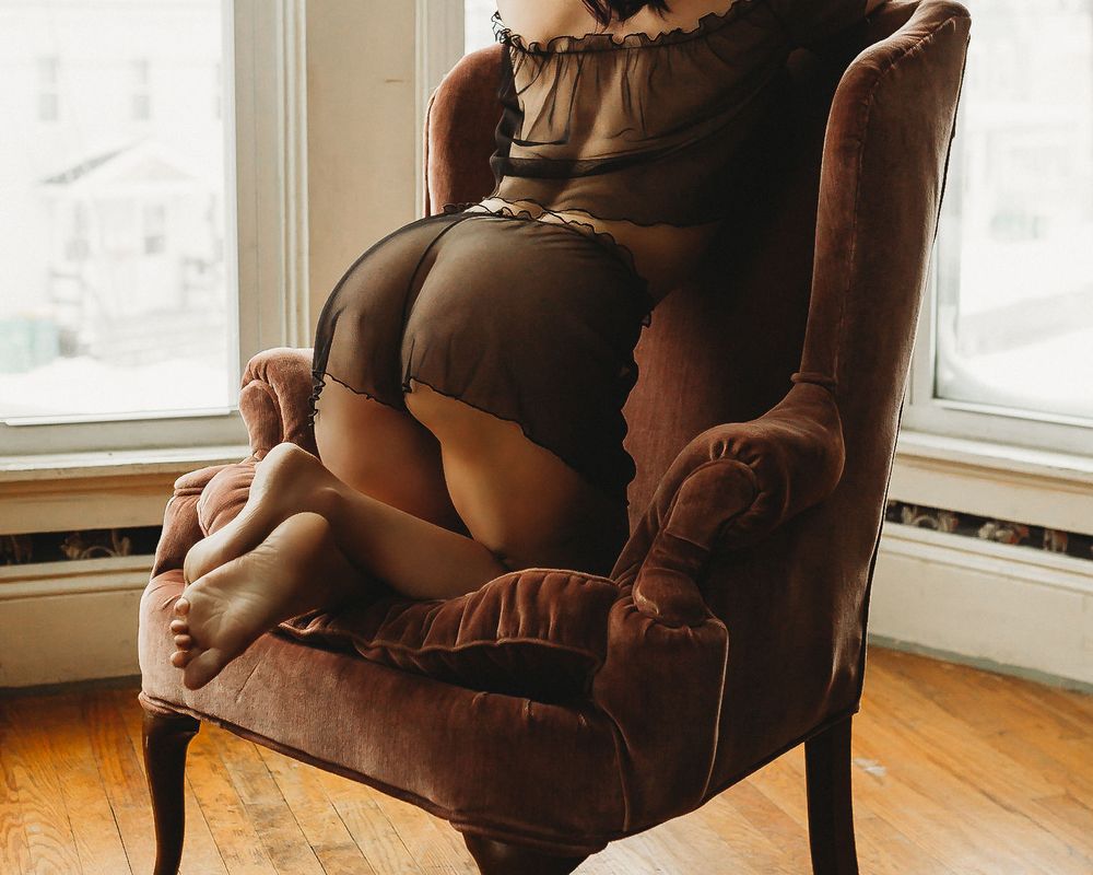 Intimate boudoir photo on velvet armchair in sunlit bay window.
