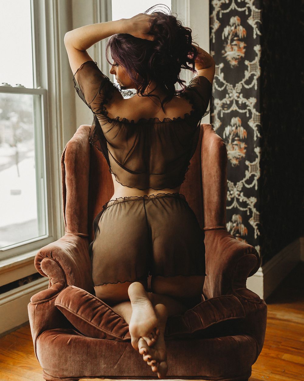 Figure poses on brown velvet chair near window with decorative wallpaper backdrop.