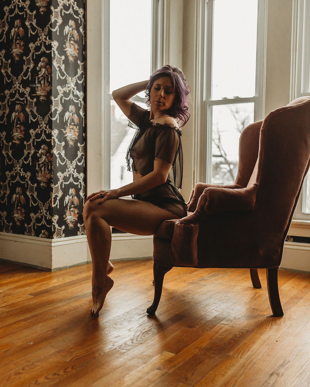 Artistic boudoir photo with brown velvet chair against patterned wallpaper.
