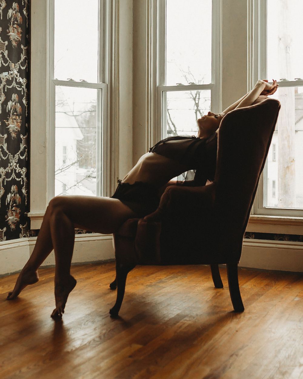 Dramatic pose in vintage armchair against bright bay windows.