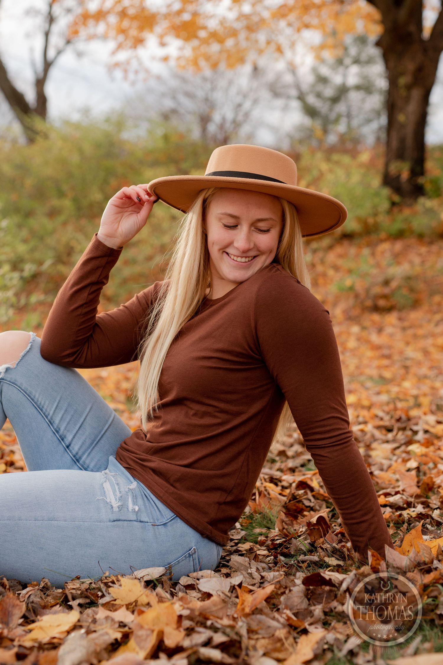 Casual fall portrait in a brown sweater and wide brim hat sitting among autumn leaves.